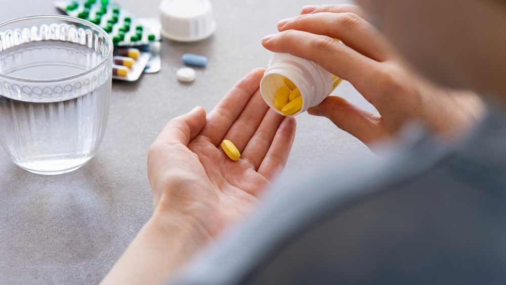 Woman holding an antibiotic tablet from a bottle with a glass of water and other blister packs on counter