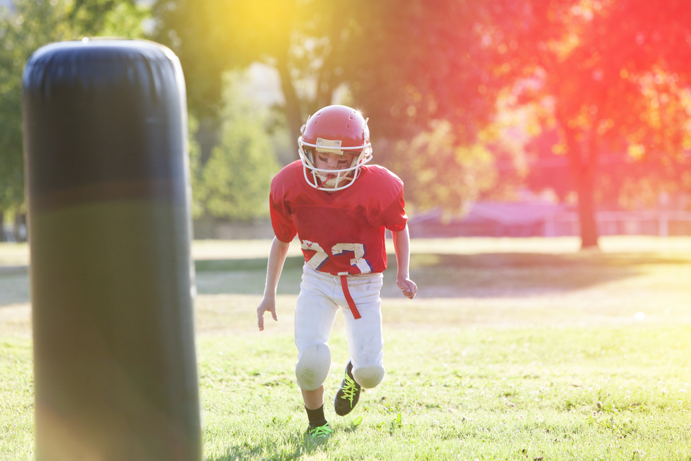 a youth football player in a red jersey and helmet running towards a tackling dummy for practice