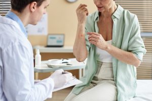 A female patient sits on an exam table with a doctor examining her skin with rash on her arm.