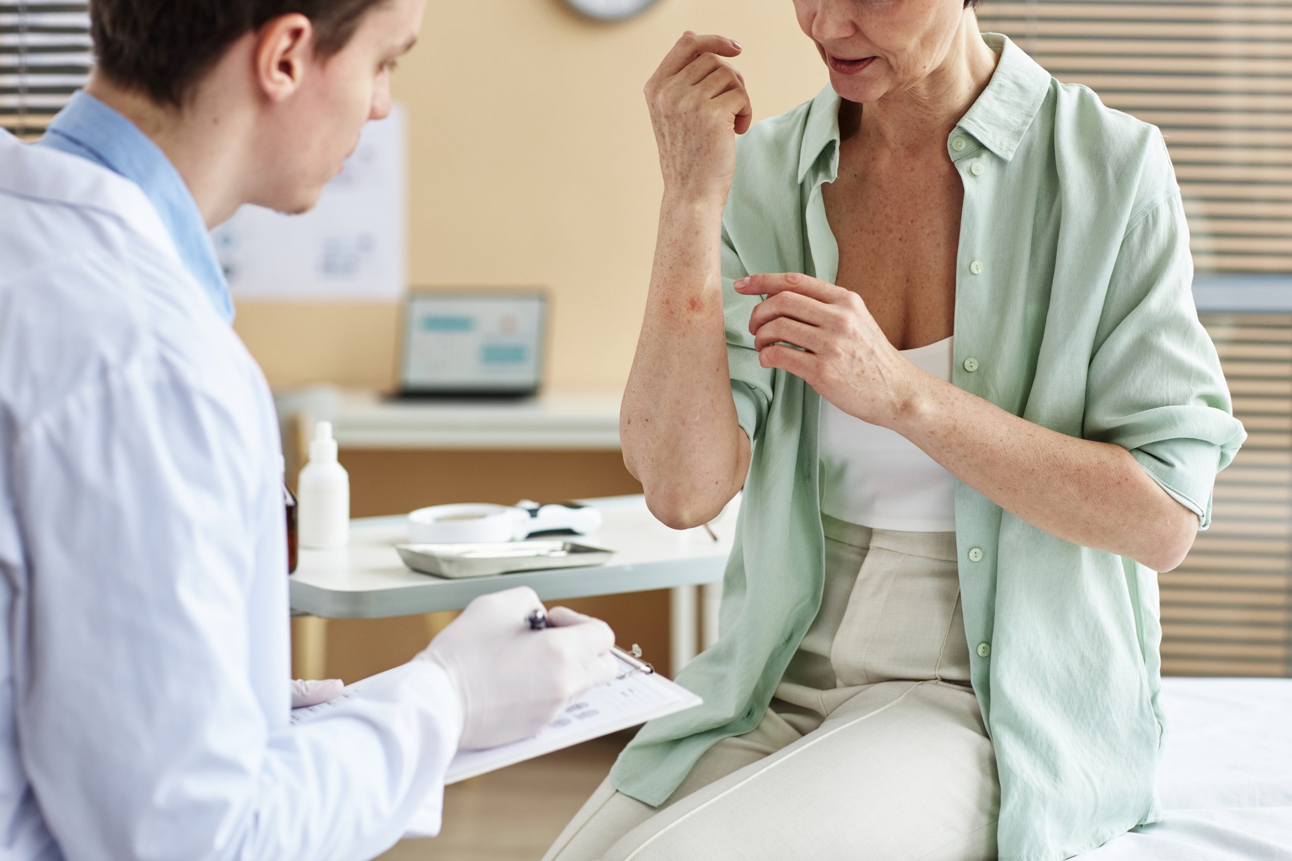 A female patient sits on an exam table with a doctor examining her skin with rash on her arm.